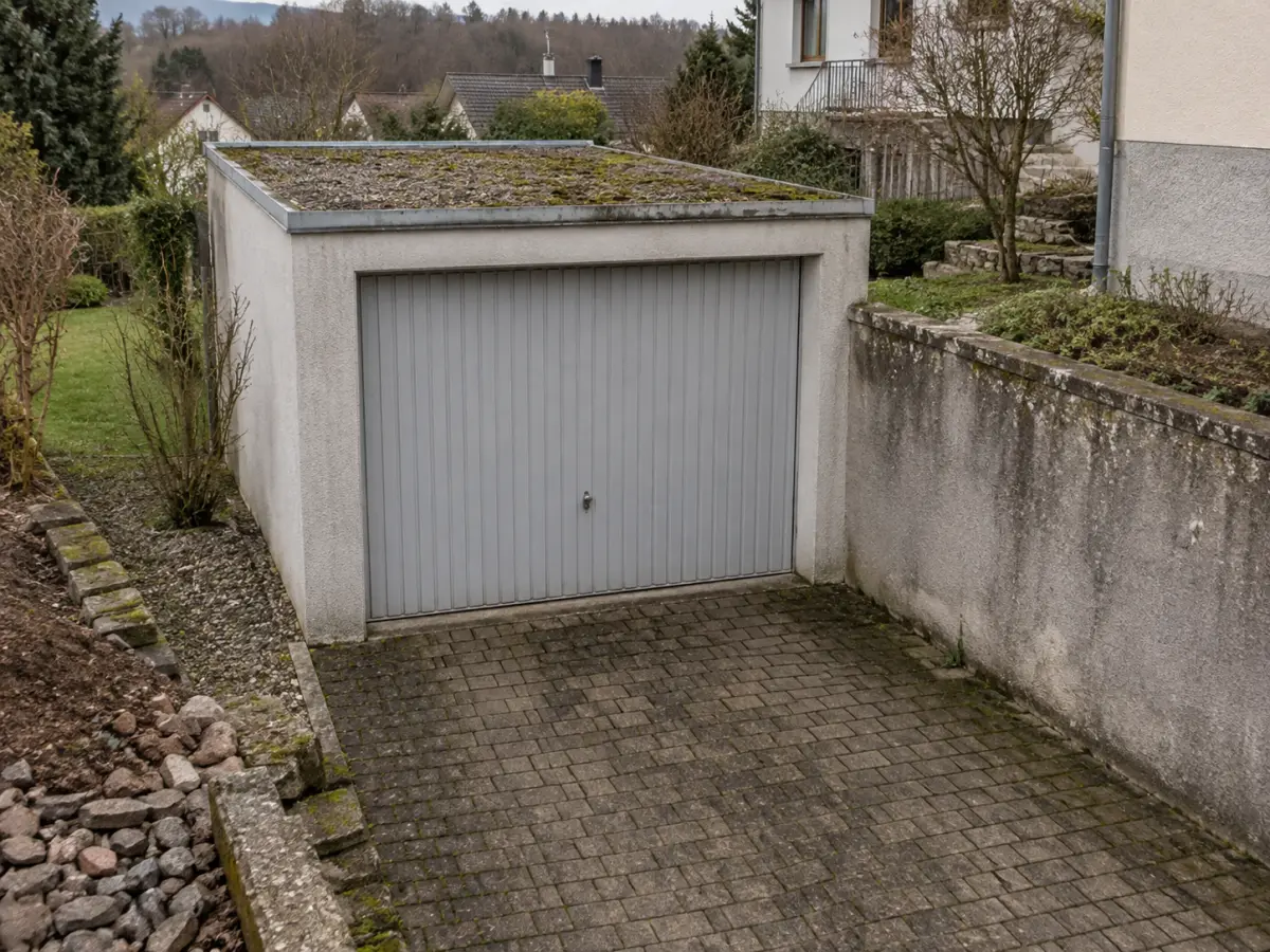 Betongarage mit geschlossener Tür, befestigter Zufahrt und angrenzender Mauer auf einem Grundstück.